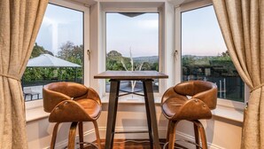 A corner room with a table and two chairs, large windows with a view of greenery and a distant landscape.