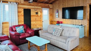 Een gezellig interieur van een chalet met een houten plafond, een aan de muur bevestigde televisie, een rode leren bank, een beige bank en een kleine houten tafel.