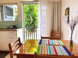 Zona pranzo con tavolo di legno, sedie, runner colorato a righe, finestra vista su vegetazione e un calendario appeso al muro.