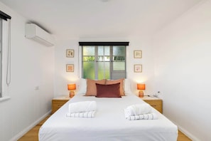 A bedroom with a bed, two bedside tables, a window with blinds, and wall-mounted air conditioning.