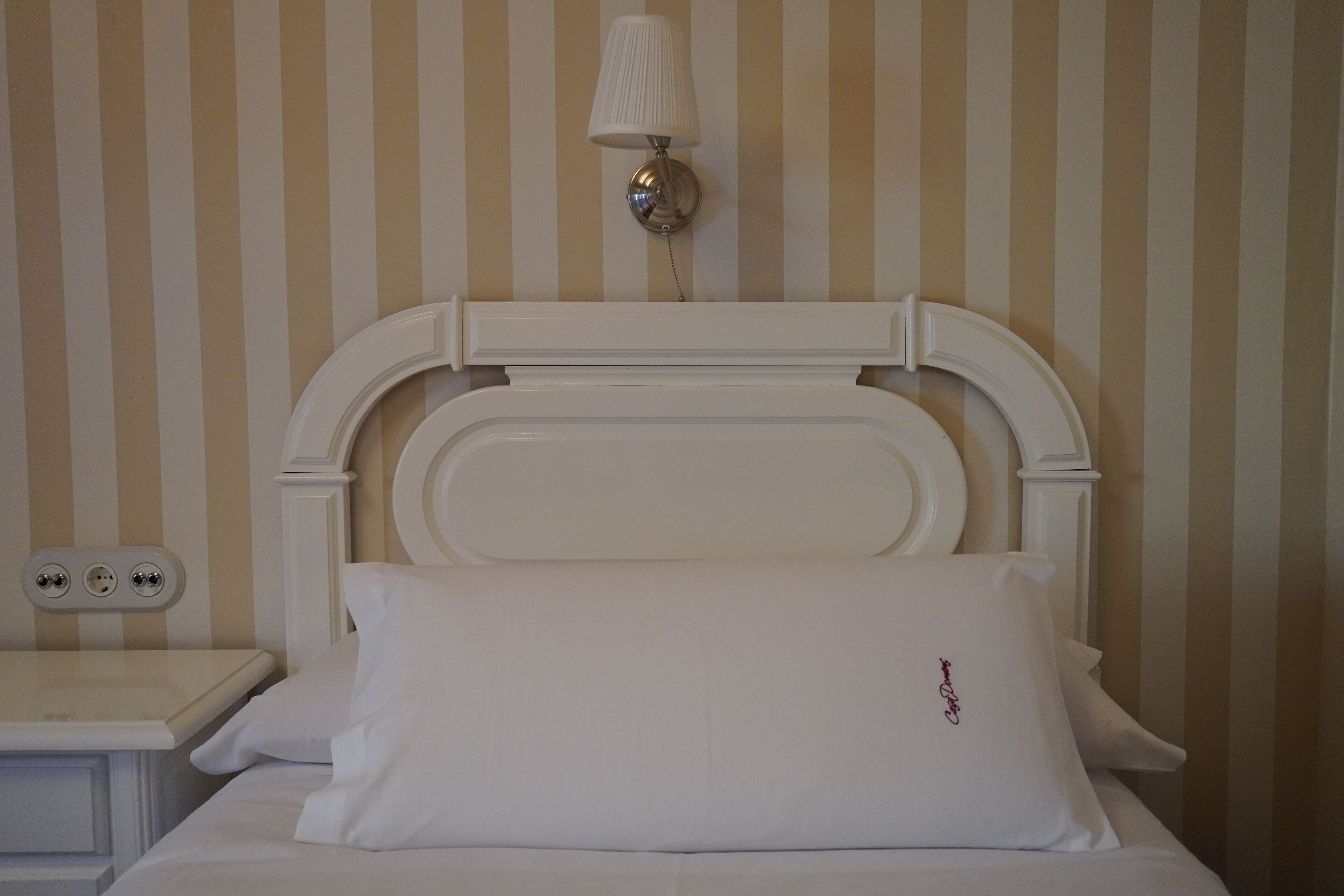 Una cama bien hecha con ropa blanca y un cabecero, una mesita de noche con una lámpara, y una pared con rayas verticales.