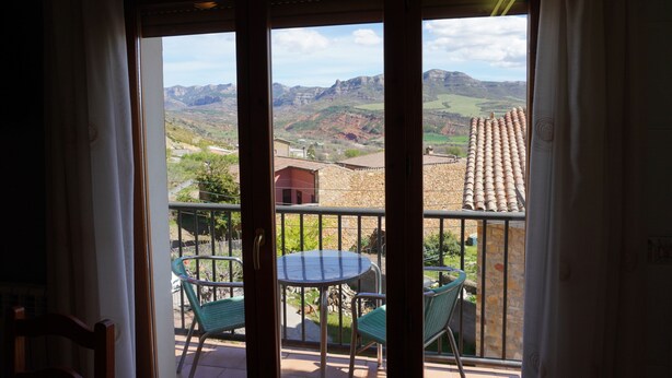 A balcony with a table and chairs, a view of mountains, and a building with a tiled roof.