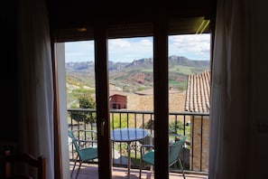 A balcony with a table and chairs, a view of mountains, and a building with a tiled roof. - Hotel Domenc (Arén)