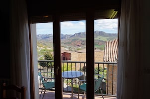 Un balcón con mesa y sillas, vista a las montañas y un edificio con tejado de baldosas.