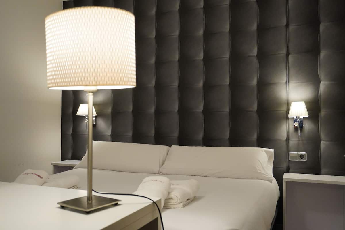 Et hotelværelse med en stor seng, to sengeborde, lamper og en moderne sengegavl.