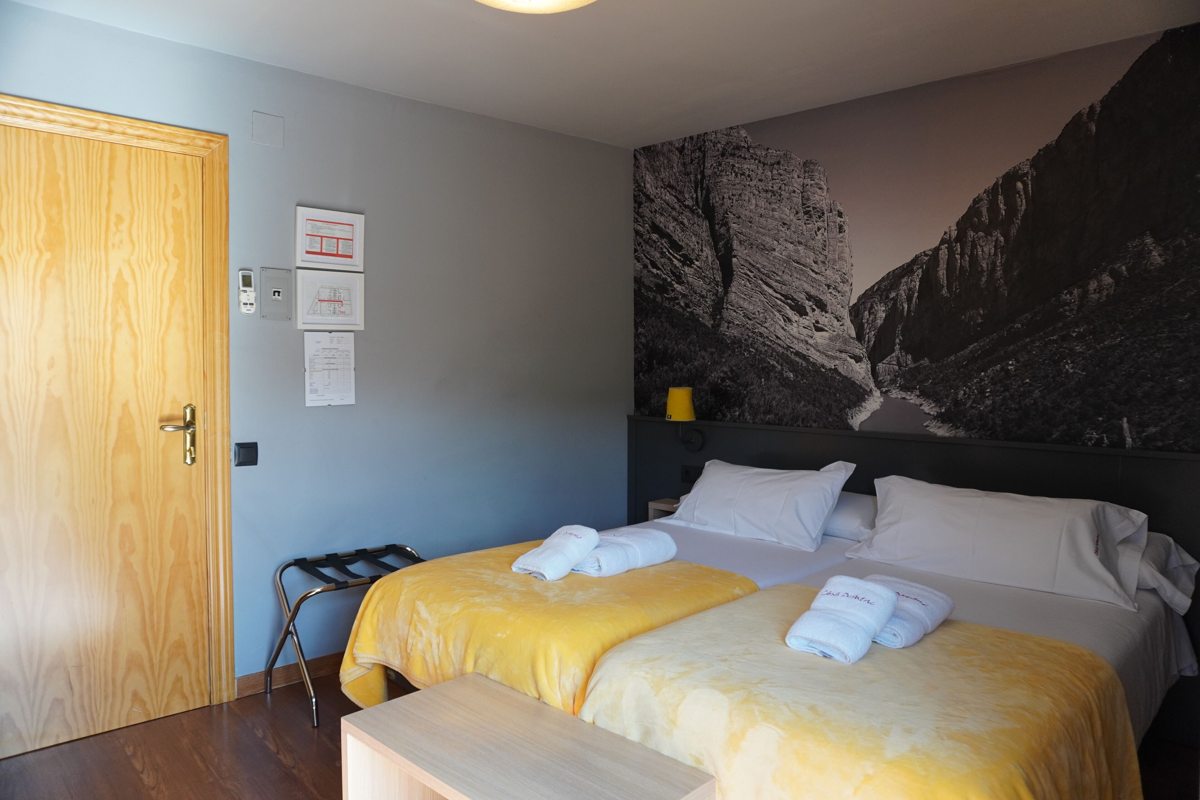 Une chambre d’hôtel avec deux lits, une porte en bois, un luminaire fixé au mur et une fresque représentant une montagne.