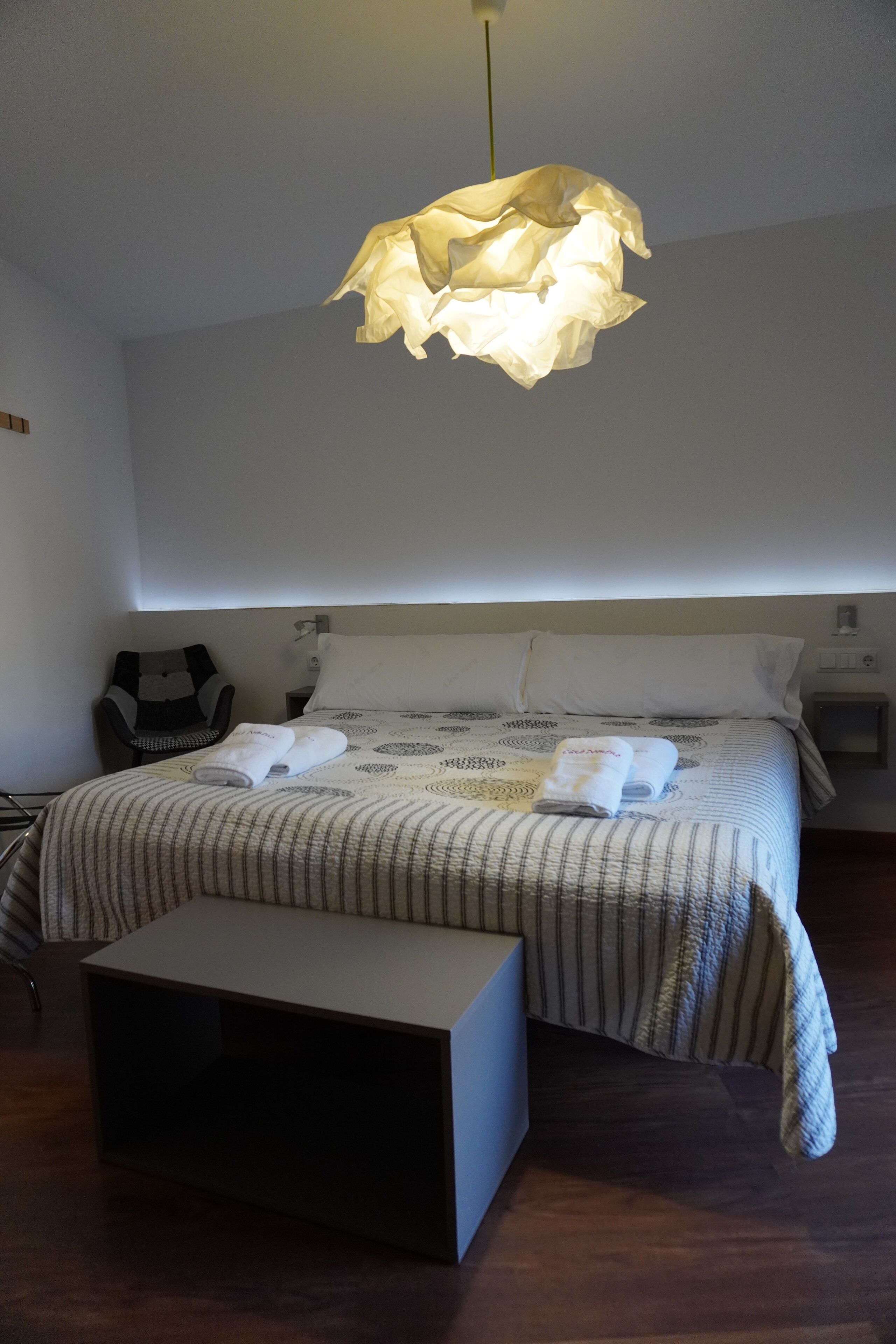 A hotel room with a bed, bedside tables, a chair, and a hanging light fixture.