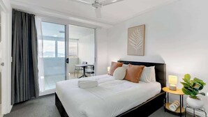 Habitación de hotel con cama, ventilador de techo, mesita de noche con lámpara, planta en maceta y puerta corrediza de vidrio que da a un balcón.