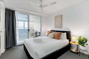 A hotel room with a bed, a ceiling fan, a bedside table with a lamp, a potted plant, and a sliding glass door leading to a balcony.