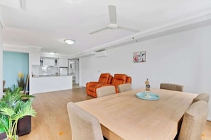 A modern living room with a dining table, orange chairs, a ceiling fan, and a kitchen in the background.