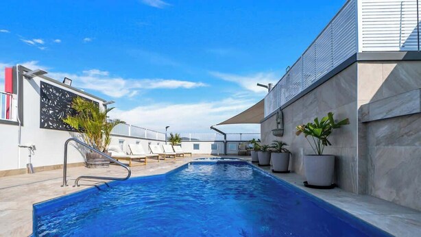 A swimming pool with a metal ladder, lounge chairs, potted plants, and a modern building with a sloped roof. - McLeod Rise—Cairn Base with Rooftop Pool & Spa (Cairns)