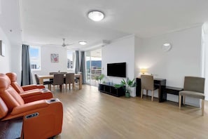 A modern living room with a flat-screen TV, massage chairs, a dining area, and a city view.