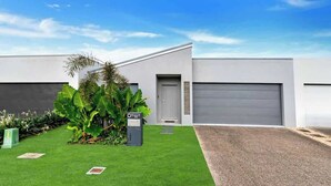 Un bâtiment moderne à la façade grise, doté d’une porte de garage et d’une pelouse bien entretenue agrémentée de plantes tropicales.