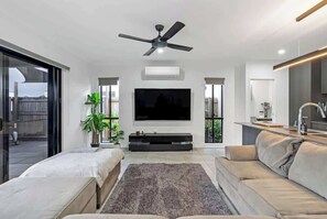 A modern living room with a flat-screen TV, ceiling fan, and a large window.