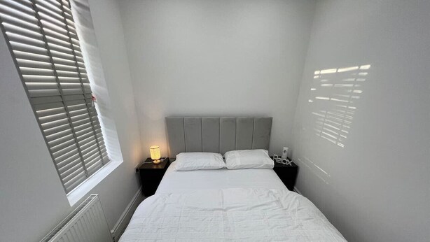 Une petite chambre à coucher, bien aménagée, avec un lit simple, une table de chevet avec une lampe et une fenêtre équipée de stores.