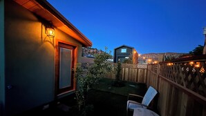Ein Garten mit einer Holzterrasse, einem Patio-Tisch und zwei Stühlen. Man sieht ein Haus mit einem Moskitonetz-Eingang und einer Laterne an der Wand.
