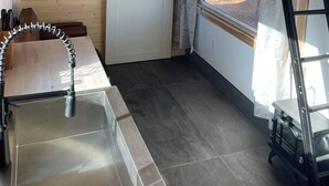 Ein kleines Zimmer mit Etagenbett, einem kleinen Tisch, einem Metallstuhl, einem Fenster mit Vorhängen und einer Leiter.