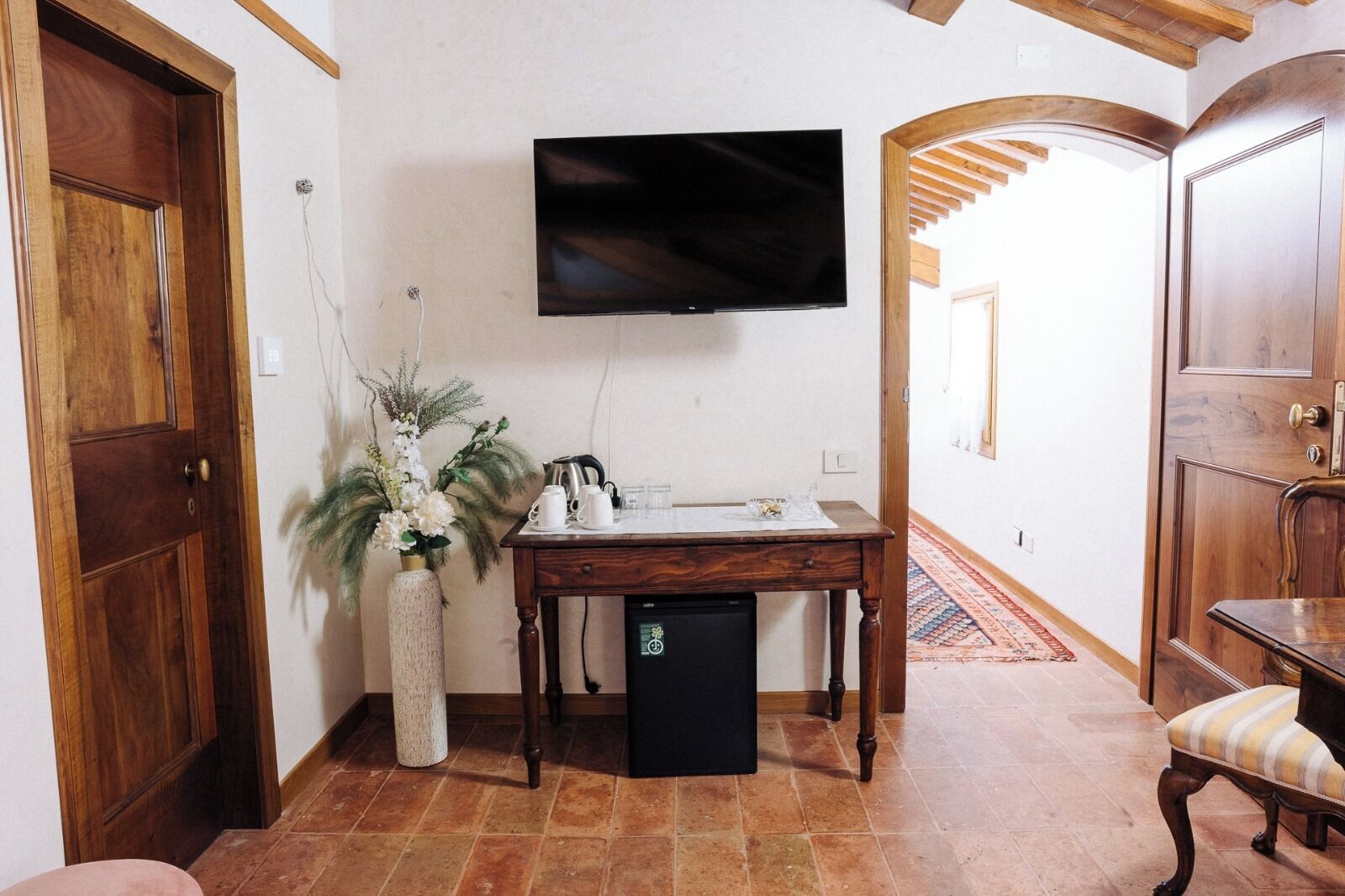 Quarto com porta de madeira, televisão na parede, mesa pequena com vaso de flores e um banco.