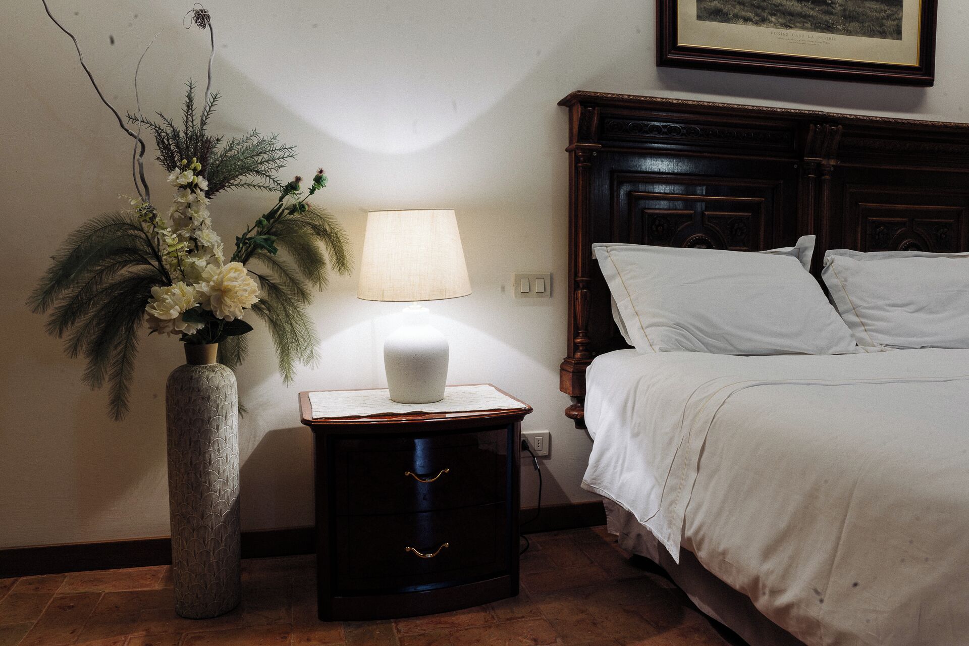A bedroom with a bed, a nightstand with a lamp, and a decorative vase with flowers.
