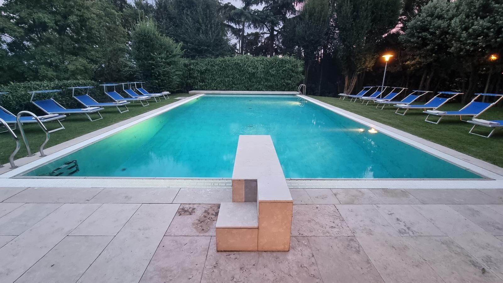 A rectangular swimming pool with a tiled deck, surrounded by blue lounge chairs and a hedge.