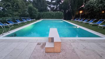 A rectangular swimming pool with a tiled deck, surrounded by blue lounge chairs and a hedge.
