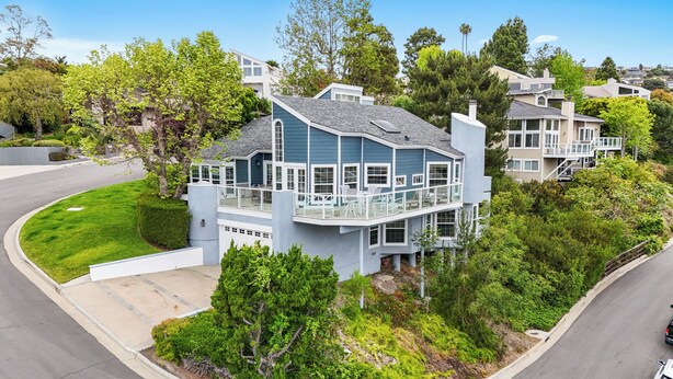 A modern two-story house with a balcony, surrounded by lush greenery and other residential buildings.