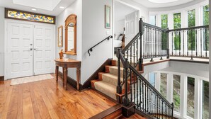 A grand staircase with a black iron railing, wooden steps, and beige carpet, leading to a balcony with white railings and a view of greenery.