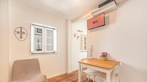 Una habitación con piso de madera, una silla blanca, una mesa de madera, una pantalla de televisión mostrando “GUEST READY” y una ventana con persianas.