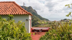 A house with a red-tiled roof, a patio with a table and chairs, and a view of a green hillside with houses. - GuestReady – Rustic Charm in Peaceful Madeira (Porto da Cruz)
