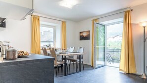 Et moderne spiseområde med et bord dækket til to, en køkkenø med hårde hvidevarer, og en balkon med gule gardiner.
