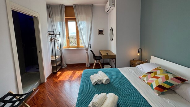 A hotel room with a bed, a desk, a chair, a window with curtains, and an air conditioning unit. - Caridei Rooms (Naples)