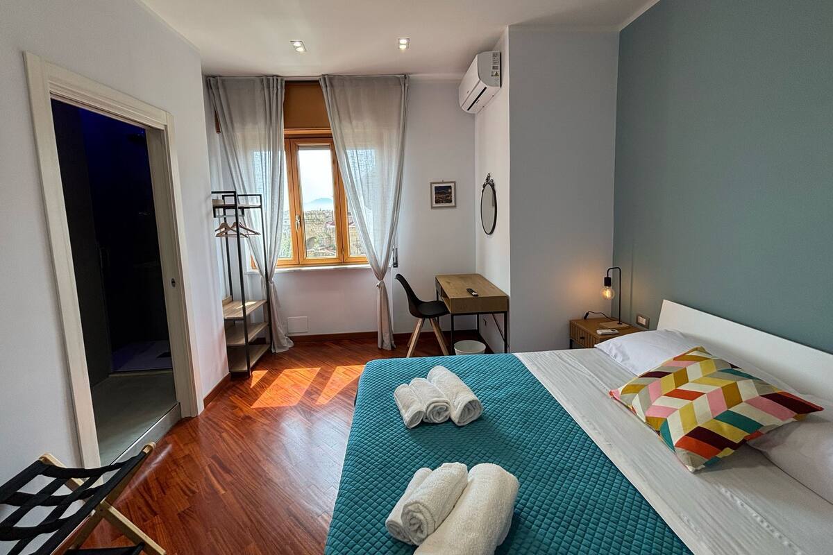 A hotel room with a bed, a desk, a chair, a window with curtains, and an air conditioning unit.