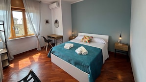A bedroom with a bed, a desk, and a chair. There is a window with curtains, an air conditioning unit, and a bedside table with a lamp. - Caridei Rooms (Naples)