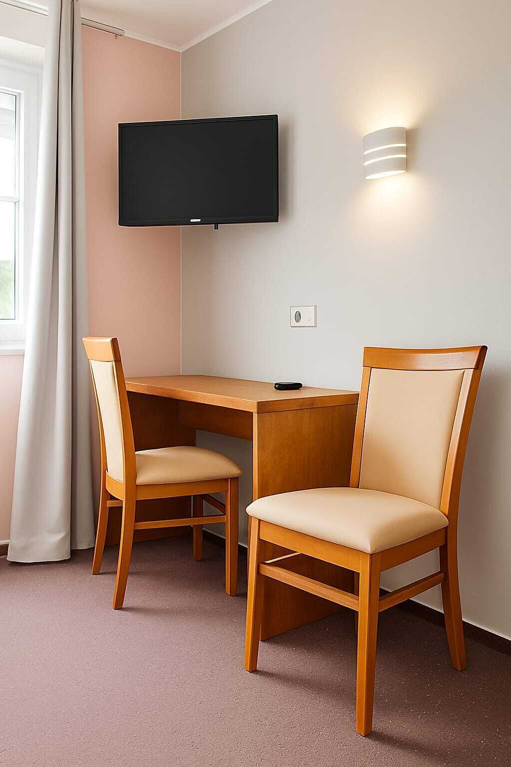 A room with a wooden desk, two chairs, a flat-screen TV mounted on the wall, and a wall-mounted light fixture.