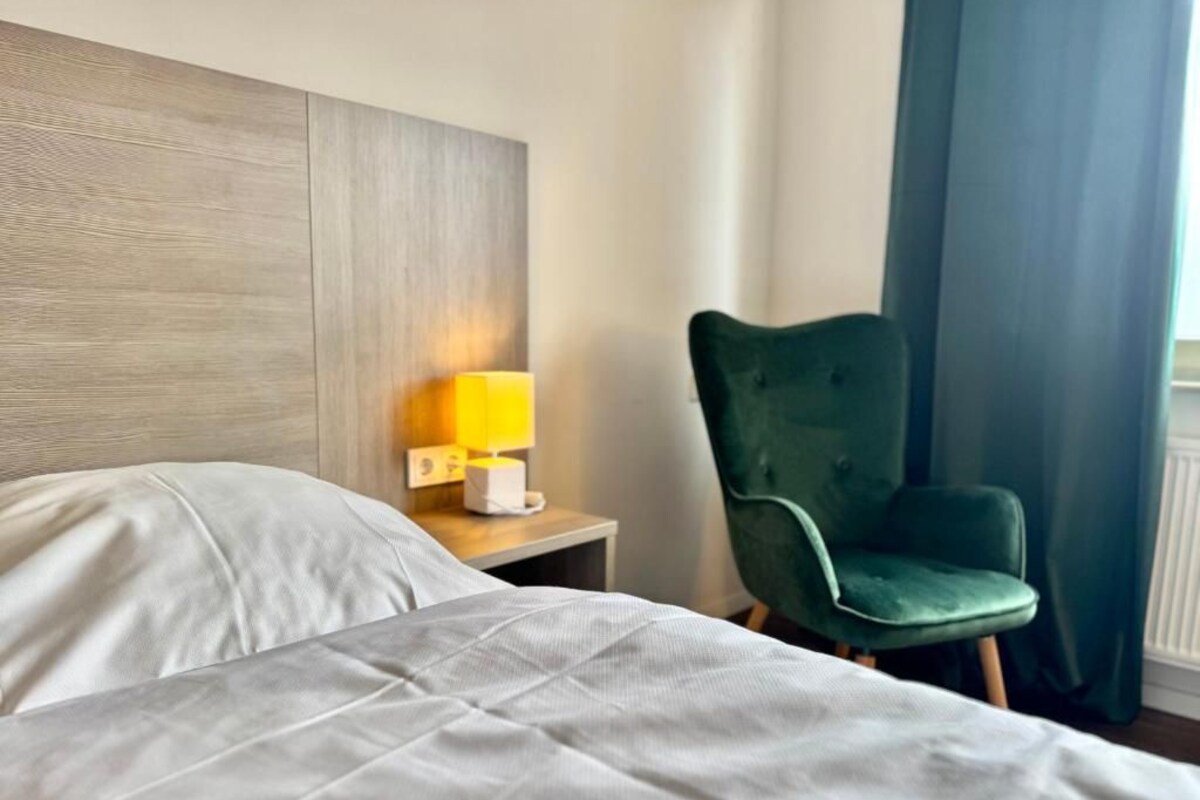A hotel room with a bed, a nightstand with a lamp, a green chair, and a window with curtains.