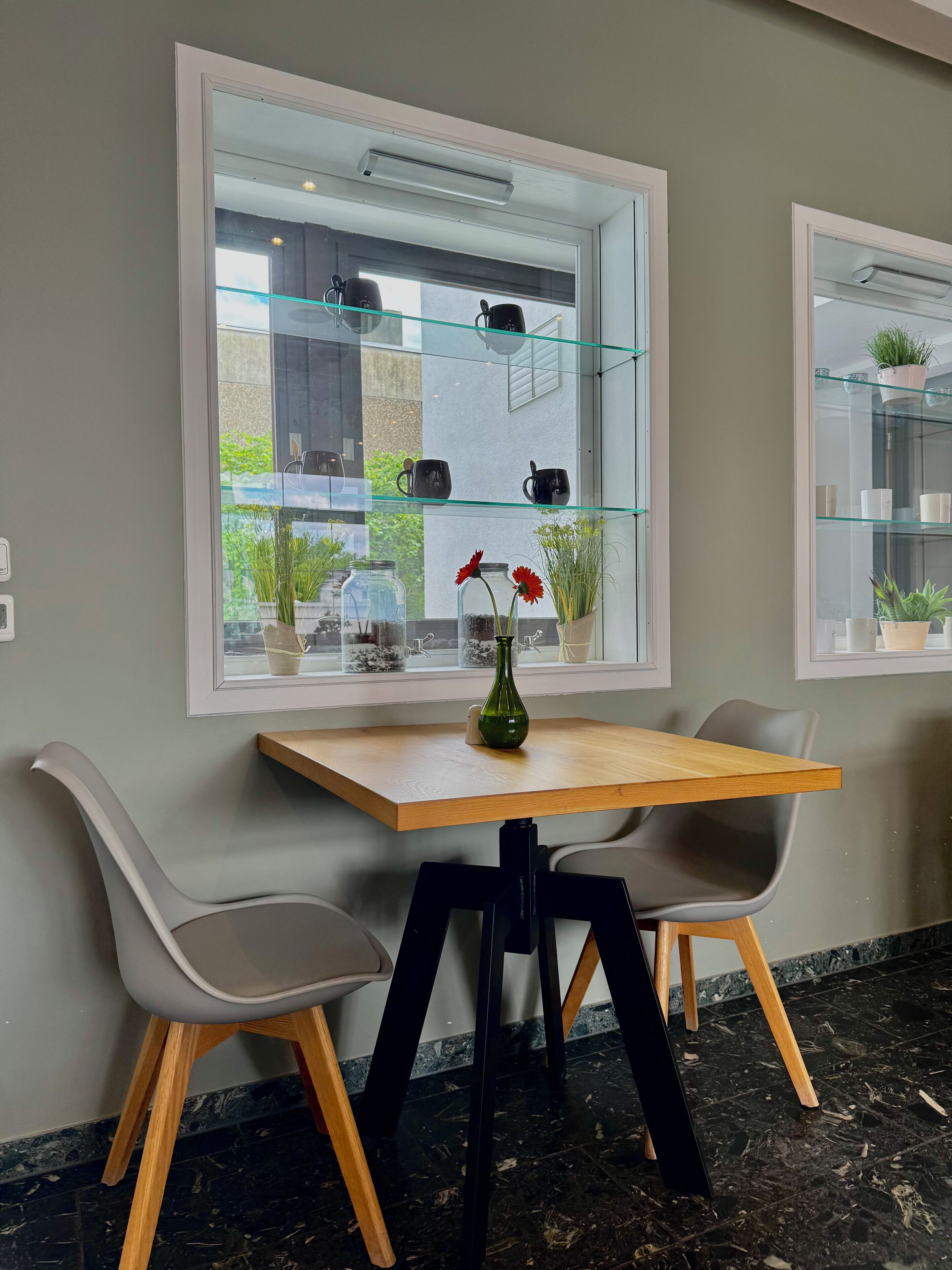 Een eethoek met een tafel en twee stoelen, een vaas met rode bloemen en een raam met planken waarop mokken en planten staan.