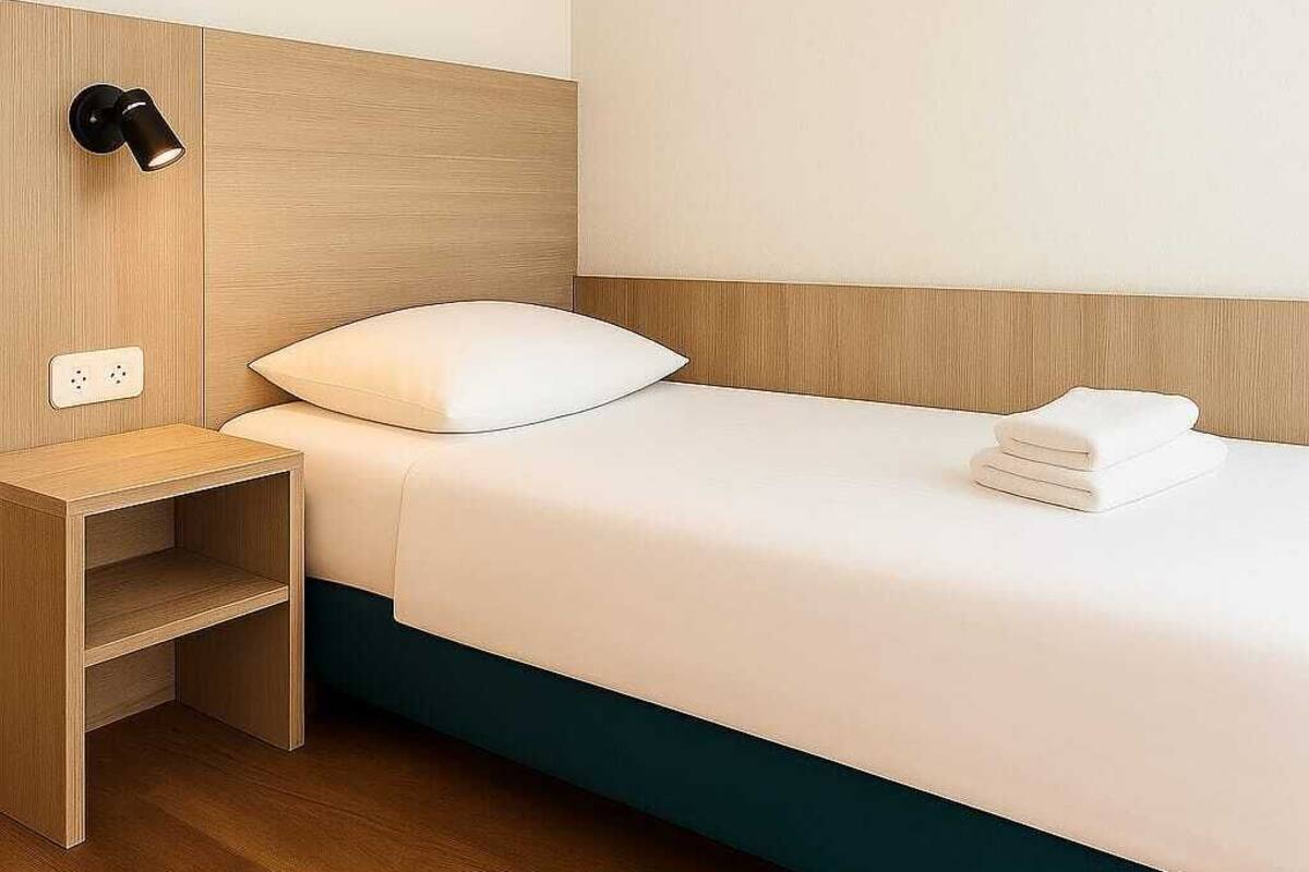 Une chambre d’hôtel avec une tête de lit en bois, un lit simple avec une literie blanche, une petite table de chevet en bois et un luminaire fixé au mur.