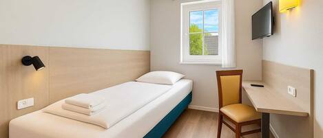 A single bed with a headboard, a desk with a chair, a TV mounted on the wall, and a window with curtains.