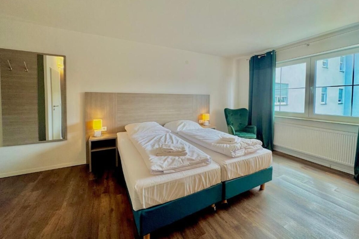 A modern hotel room with a large bed, bedside lamps, a green armchair, and a view of buildings through the window.