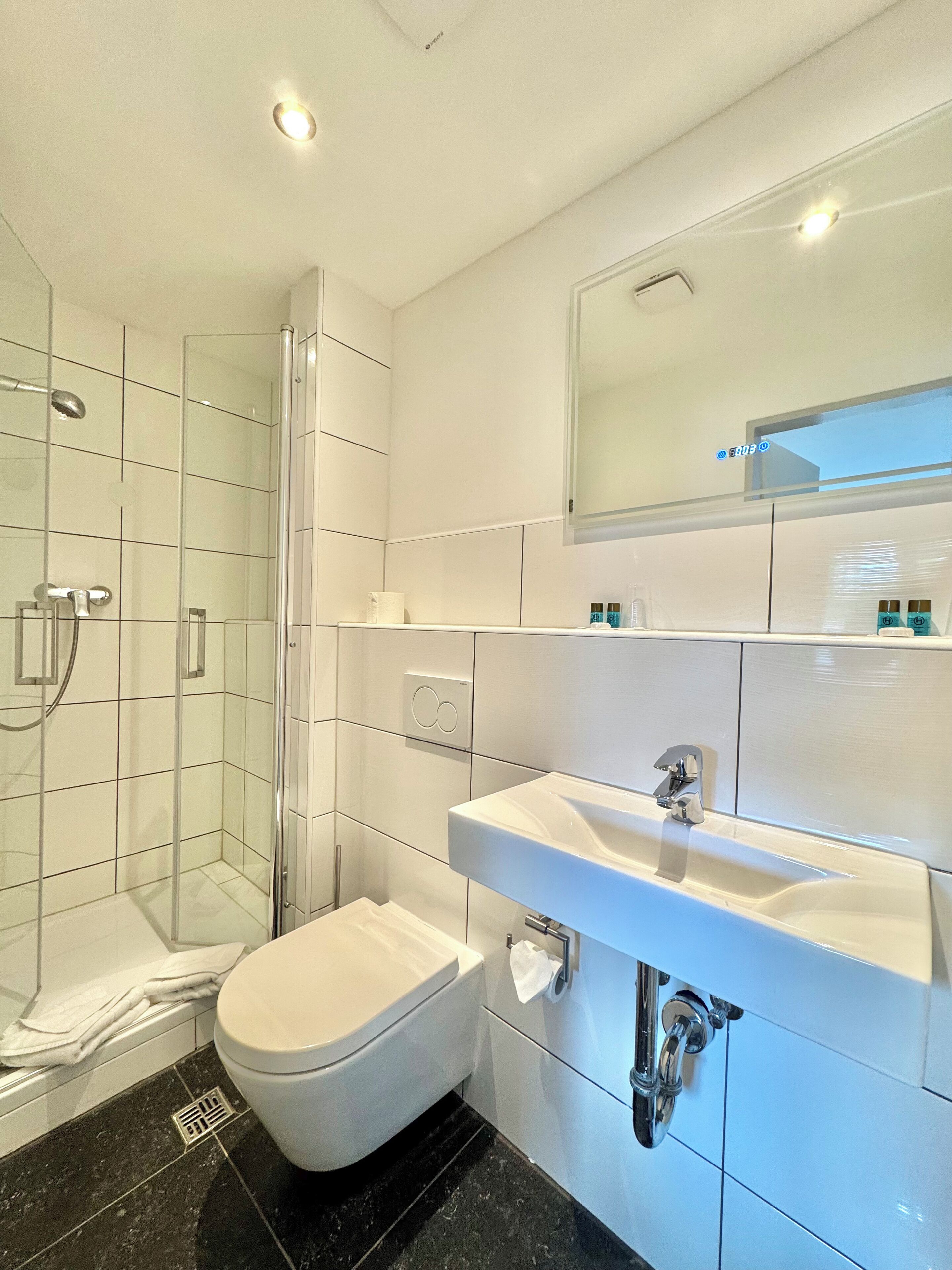 Un baño moderno con cabina de ducha de vidrio, inodoro blanco y un espejo grande sobre el lavamanos.