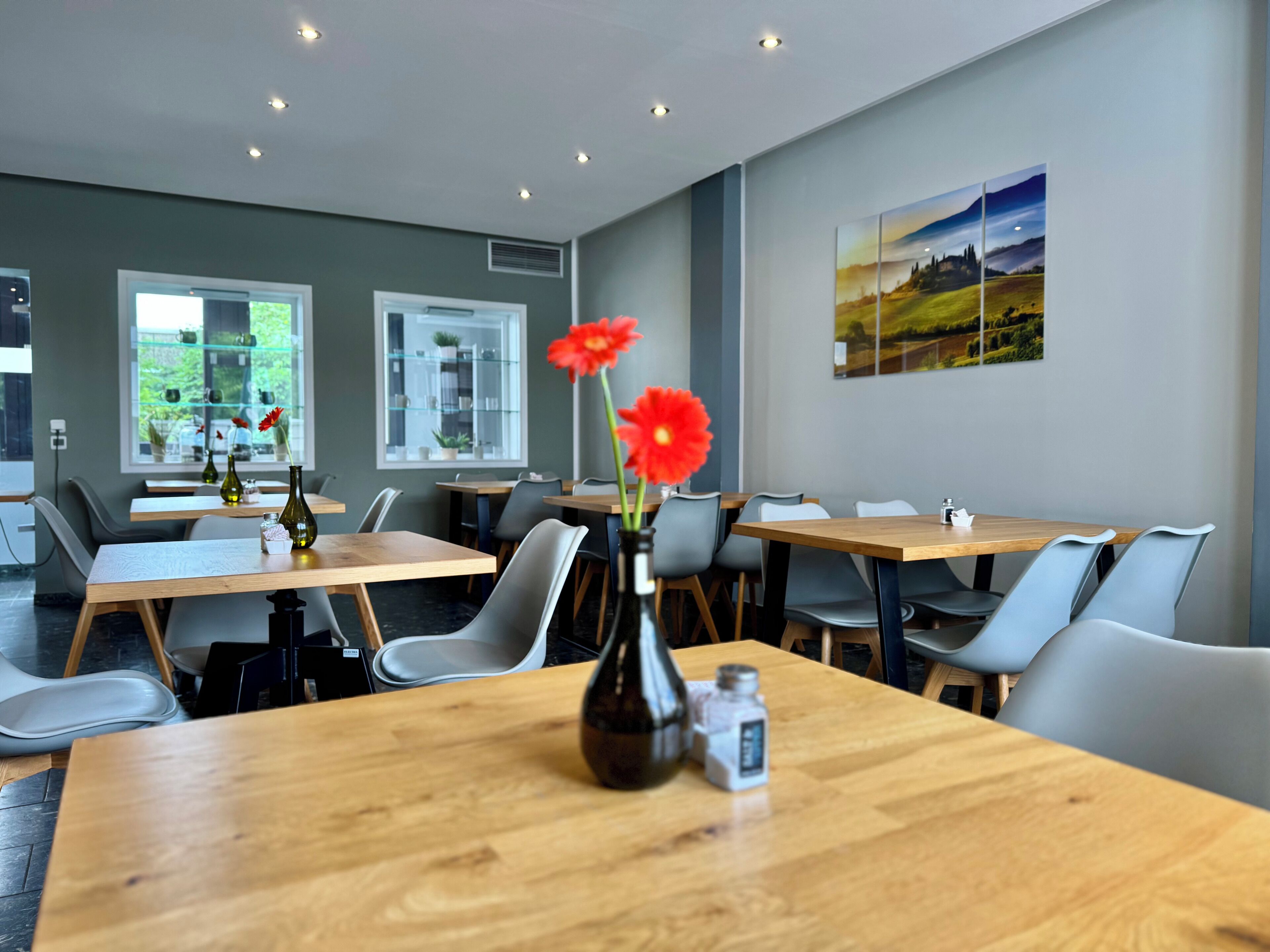 Een moderne eetruimte met houten tafels en grijze stoelen, met een vaas met rode bloemen en een zoutvaatje op tafel.