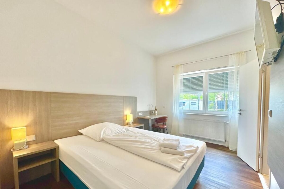A hotel room with a bed, bedside tables, a desk, and a window with blinds.
