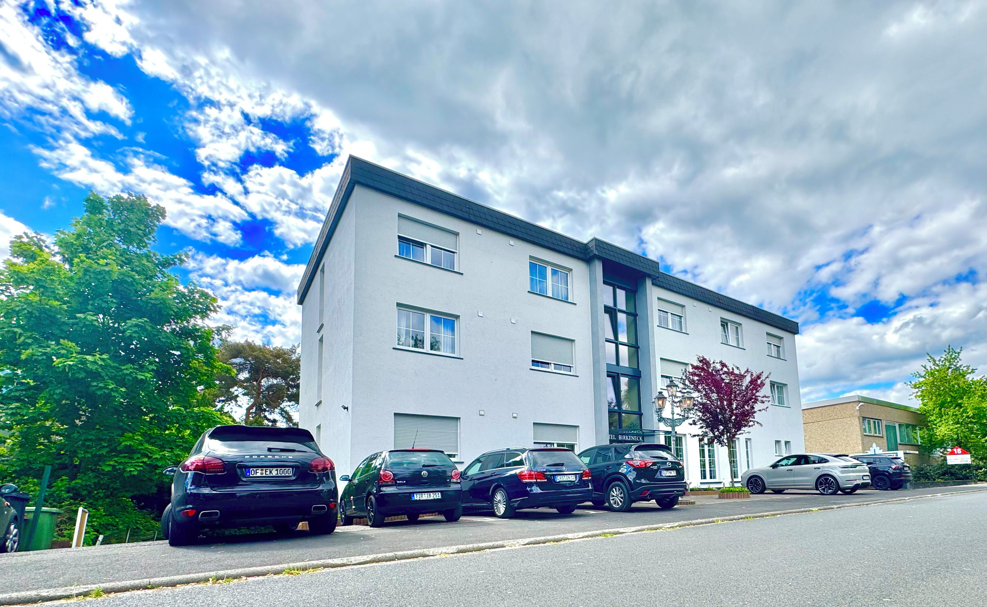 A modern building with multiple floors, parked cars in front, and a clear sky with clouds.