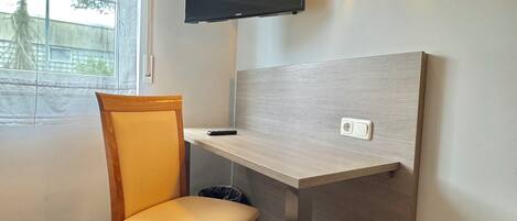 Una habitación pequeña y moderna con un escritorio de madera, una silla, un televisor montado y una ventana con vistas a árboles.