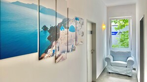 Um corredor com uma grande obra de arte na parede retratando uma paisagem costeira, uma porta branca e uma poltrona branca perto de uma janela com vista para vegetação.