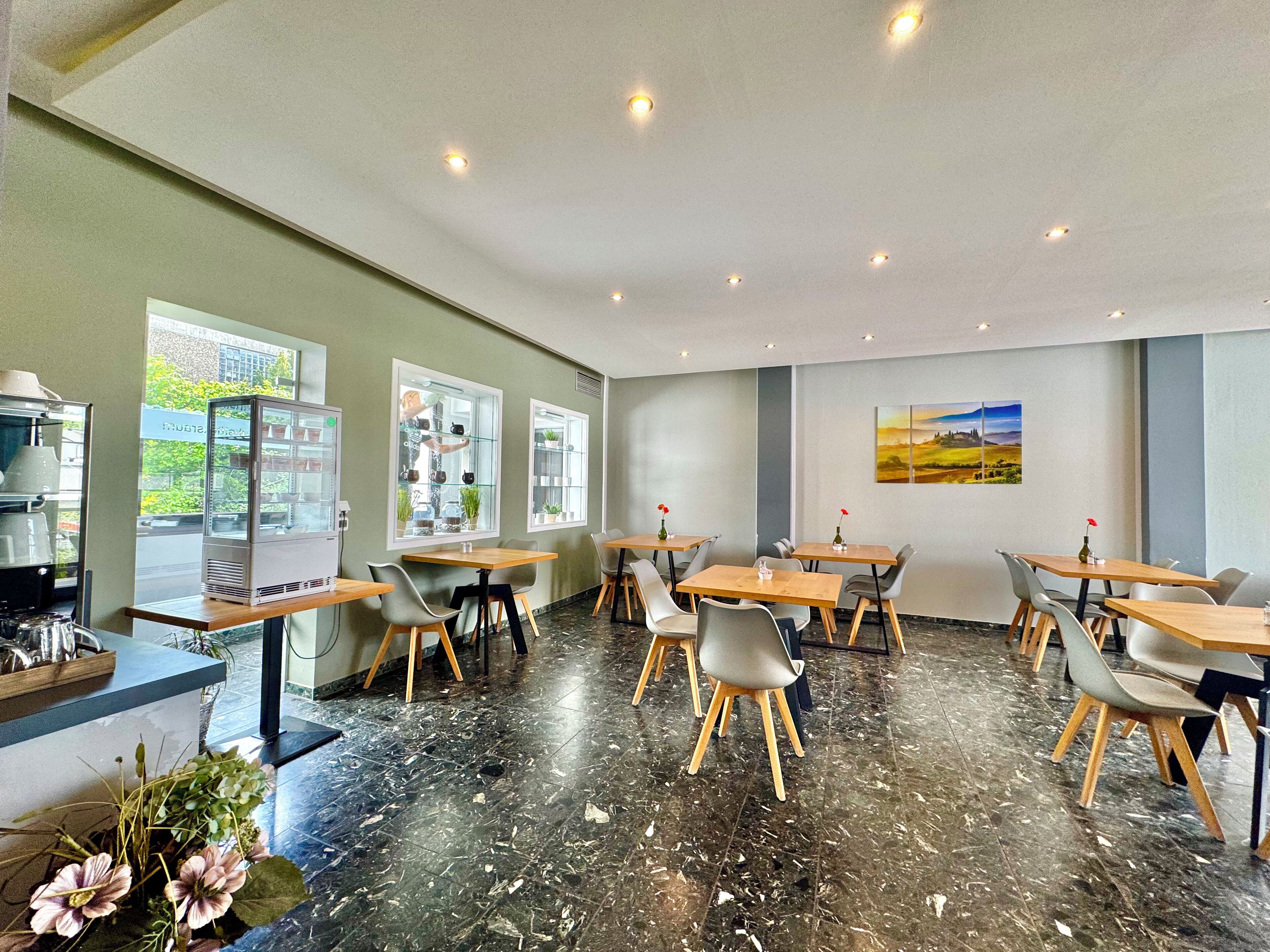 Una cafetería moderna con suelo de mármol, mesas y sillas de madera, y grandes ventanales con vistas a zonas verdes.