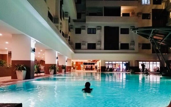 An indoor swimming pool with a person swimming, surrounded by a multi-level building with balconies and air conditioning units.