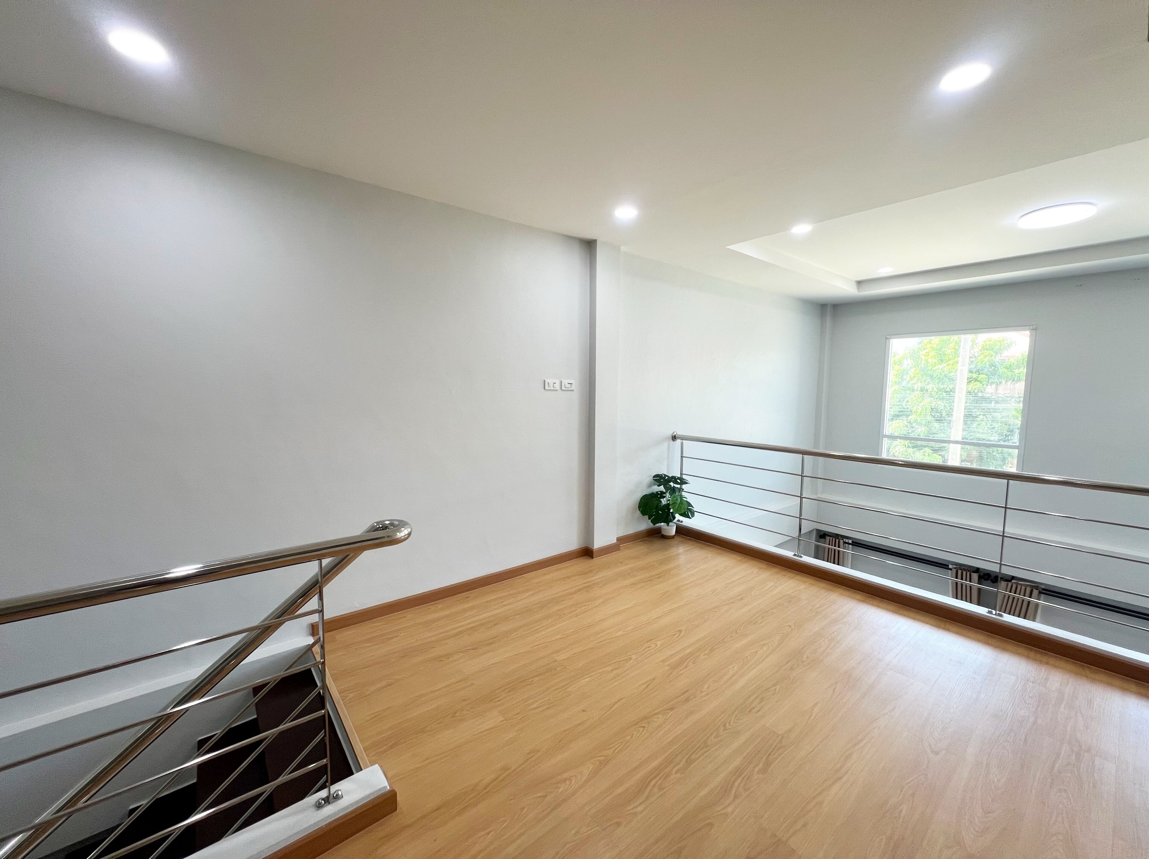 A spacious room with a wooden floor, a metal railing, and a window with a view of greenery.