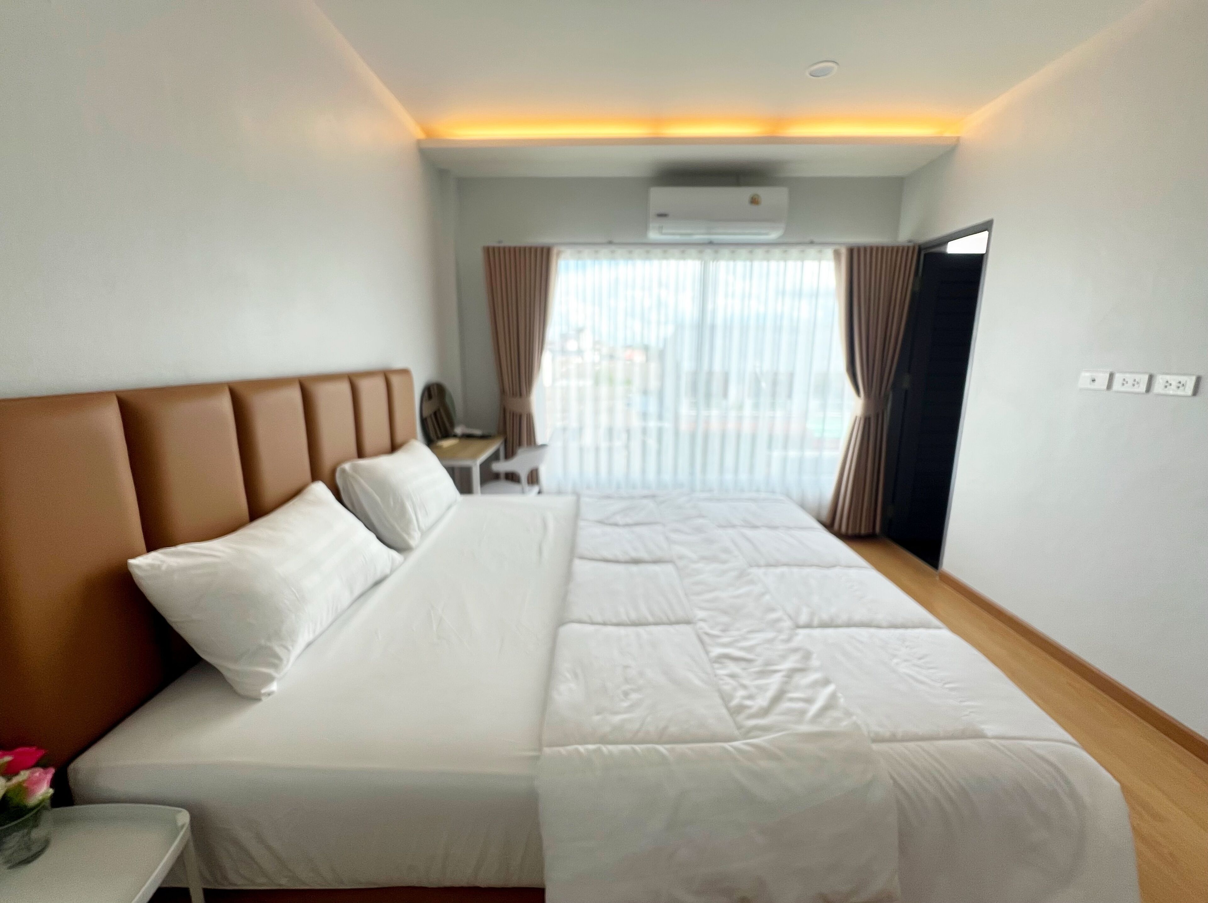 A hotel room with a large bed, a desk, and a chair. There is an air conditioning unit on the wall and a window with curtains.