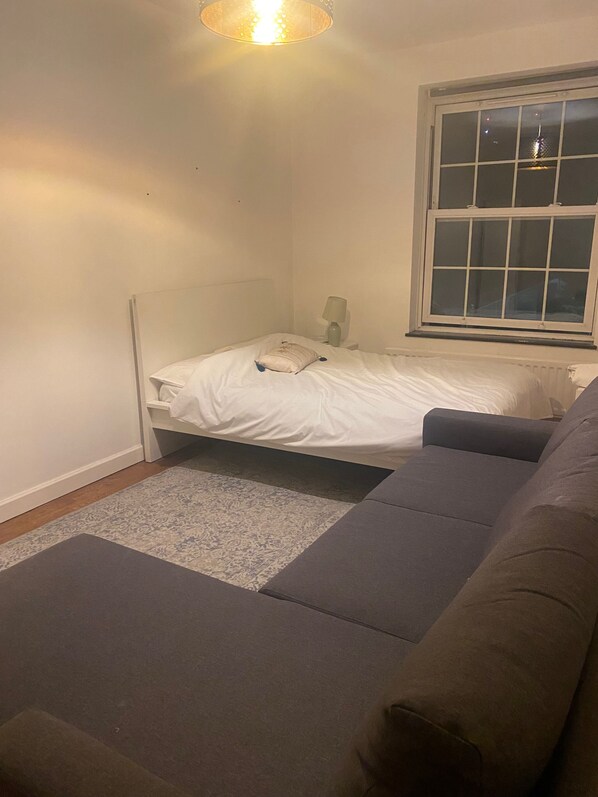 A bedroom with a bed, a nightstand, a lamp, a window, and a gray sofa. - 4 Bed Flat - Sleeps 10 - Pets - Parking (London)
